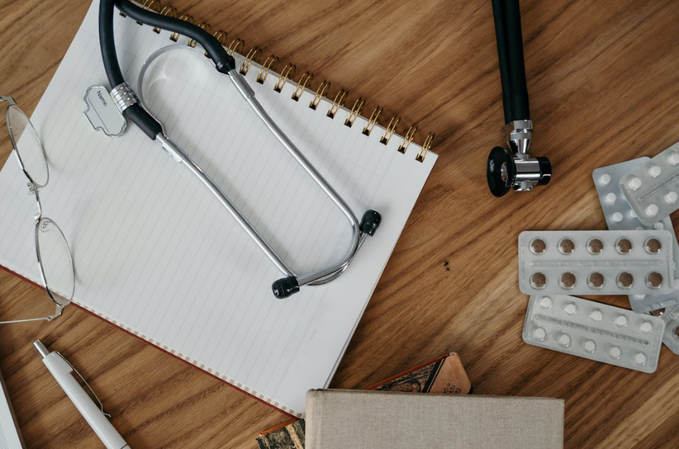 Stethoscope, glasses, notebook, and blister packs of medication on a wooden desk