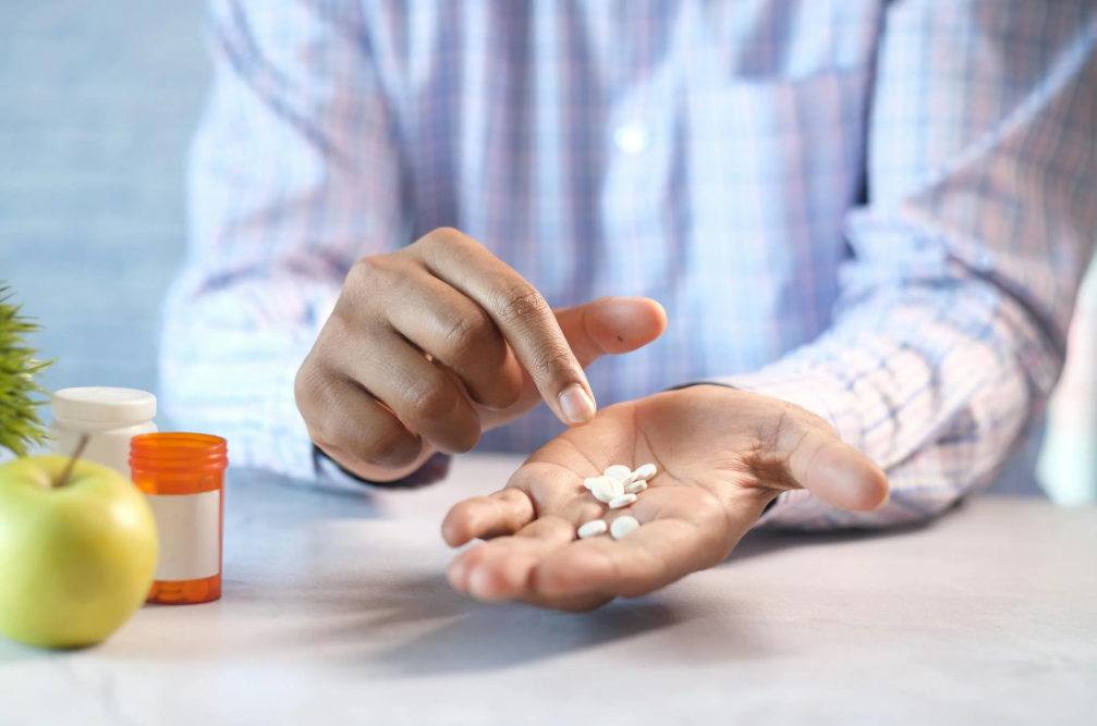 Hand holding white pills with bottles and an apple on the table.