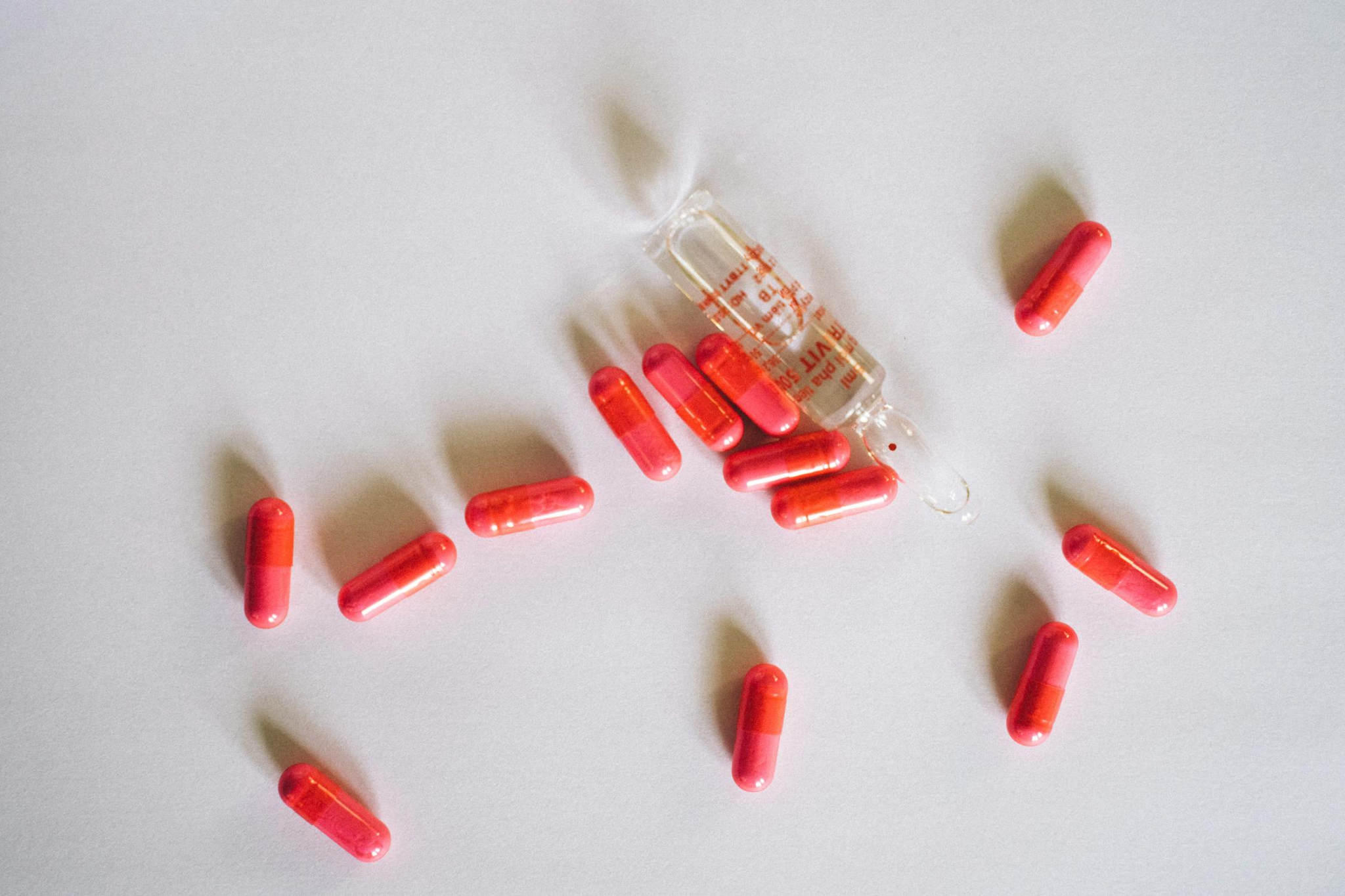 A glass ampoule and numerous red capsules scattered on a white surface.