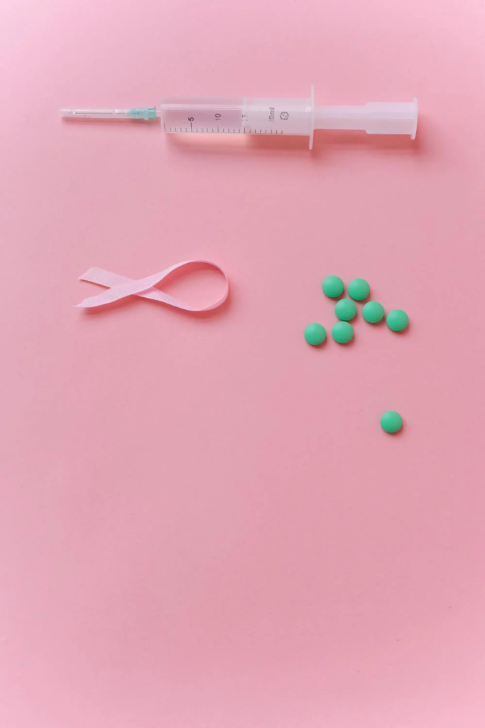 A syringe, a pink awareness ribbon, and green pills on a pink background.